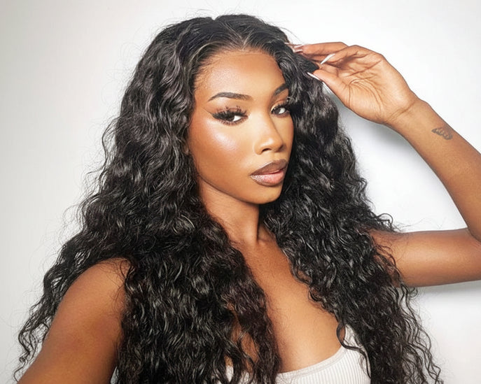 Woman with long, wavy black hair styled in a wig, wearing a white top against a light gray background.